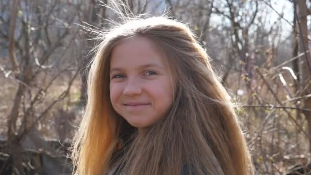 Joyful blonde girl looking into camera and laughing outdoor. Portrait of female smiling child against the blurred background of a forest at early spring. Positive emotions of little kid on her face - Footage, Video
