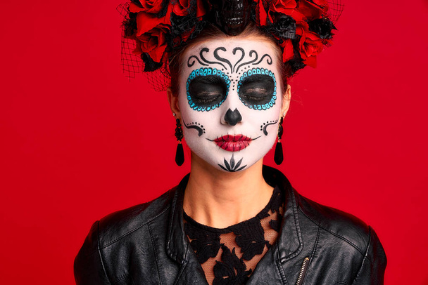 Τρομακτική νεαρή ανατριχιαστική κυρία Καλαβέρα. φοράει καλλιτεχνικό make-up για τη γιορτή όλων των νεκρών.Φοράει μαύρο δερμάτινο μπουφάν και δαντελένια γάντια, ντυμένο σκελετό απομονωμένο με κόκκινο - Φωτογραφία, εικόνα