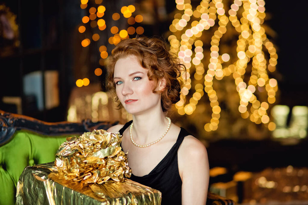 クリスマスギフトボックスを持つ若い女性は、金の装飾が施されたクリスマスツリーの背景に贈り物を保持しています。スタイリッシュな女性の、ファッショナブルなイブニングウェアとアクセサリー、メイク。明けましておめでとうございます. - 写真・画像
