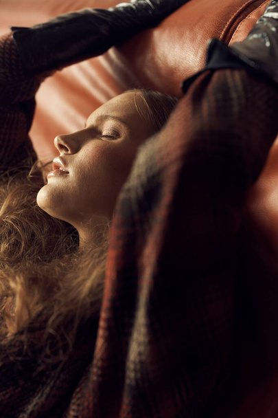 portrait photo of a girl with brown curly tails in a vintage checkered jacket, she is sitting on a leather sofa, her arms raised and her eyes closed - Foto, Imagen