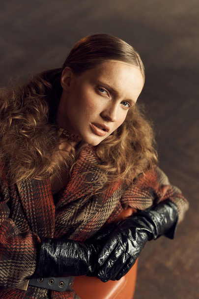 portrait photo of a girl with freckles, brown curly tails in a vintage checkered jacket, she mysteriously looks into the camera sitting on a leather chair - Photo, image