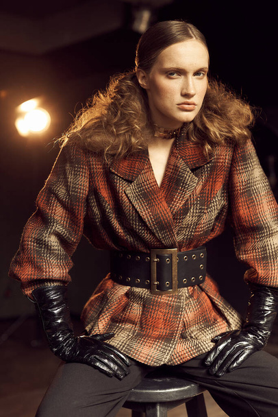 stylish curly girl in a vintage checkered jacket, leather gloves and black pants sits on a chair in the loft studio and looks into the camera, behind her shining lantern - Фото, зображення