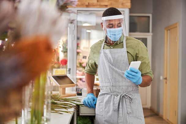 Young florist using smartphone during pandemic at work - 写真・画像