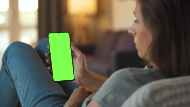 Žena doma leží na pohovce a používá smartphone se zelenou maketou obrazovky ve vertikálním režimu. Dívka prohlížení Internetu, sledování obsahu, videa, blogy. - Záběry, video