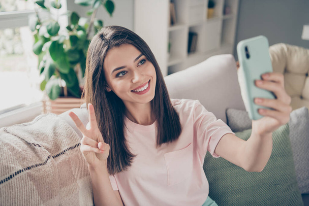 Photo of homey lady hold telephone make selfies show v-sign symbol sit sofa indoors - Foto, imagen