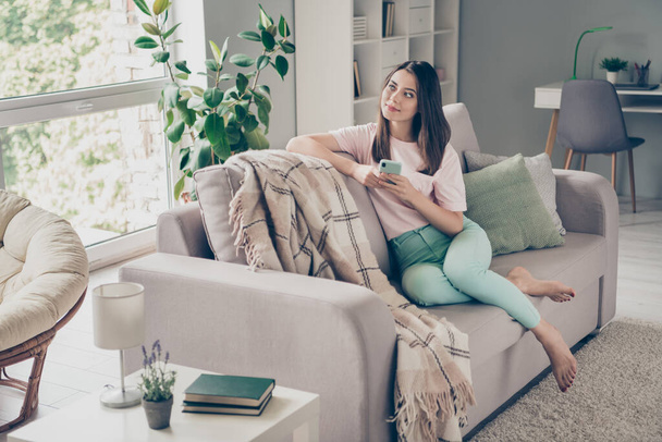Photo portrait of dreamy woman looking into the window sitting on couch holding phone in two hands indoors - Foto, afbeelding