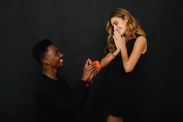 A young black man gives his Caucasian girlfriend a Christmas or Valentines Day gift and proposes to her while standing on one knee - Foto, Imagem