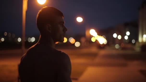Um jovem homem musculoso sem uma camiseta fica contra o fundo de luzes embaçadas da cidade noturna e se vira e olha para a câmera. Vista traseira. Close-up - Filmagem, Vídeo