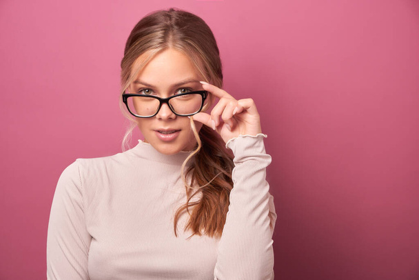 Portrait of a beautiful girl with glasses looking at the camera lens. Girl holding sexy glasses with her fingers in gray clothes on a pink background - Fotó, kép