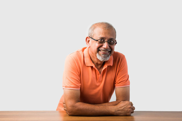 Porträt eines indischen asiatischen Seniors, der am Tisch sitzt und präsentiert oder in Erfolgspose posiert - Foto, Bild