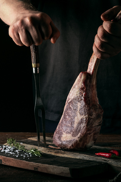 Ein Mann hält ein rohes Tomahawk-Steak auf einem Knochen aus getrocknetem Rindfleisch vor dunklem Hintergrund - Foto, Bild