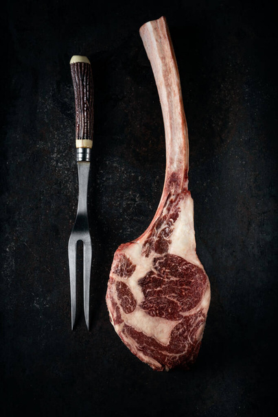Rauwe Tomahawk biefstuk droog gerijpt met een vleesvork op een donkere stenen achtergrond, bovenaanzicht - Foto, afbeelding