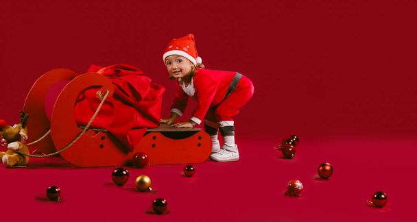 Christmas, family holiday. Children are waiting for gifts and magic. The little girl is happy that she found a sleigh and a sack of Santa Claus. Photo on a red background - Zdjęcie, obraz