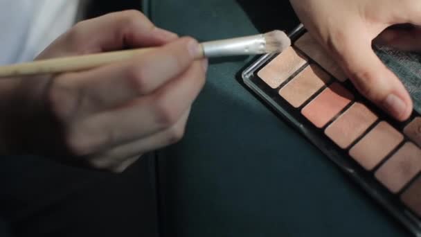 A young woman uses a palette of pastel eyeshadows to apply makeup and holds a cosmetic brush in her hand. Close-up of hands - Footage, Video