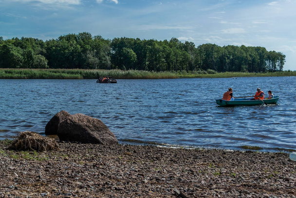 Pequeños botes de remo de pasajeros con la familia, la gente flota en el lago en el parque entre árboles verdes, arbustos, bosques en tiempo soleado. Piedras costa. Rusia verano - Foto, Imagen