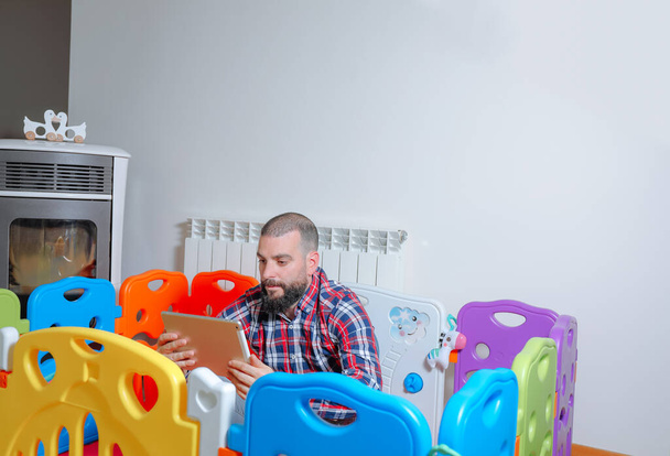 Padre joven sentado dentro del parque infantil usando una tableta digital en casa. Concepto de serie online - Foto, Imagen