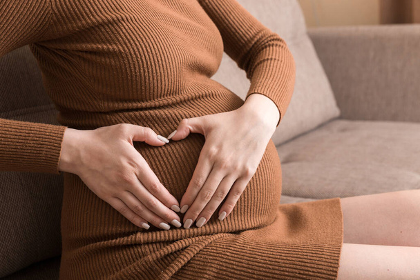 Schnappschuss von schwangerer Frau, die Bauch berührt und Handherz-Symbol auf Sofa zu Hause zeigt. - Foto, Bild
