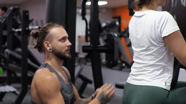 Μάθημα γυμναστικής με ένα νεαρό αστείο εκπαιδευτή μια αρχάρια γυναίκα κουνάει την πλάτη και τα χέρια της στον προσομοιωτή - Πλάνα, βίντεο