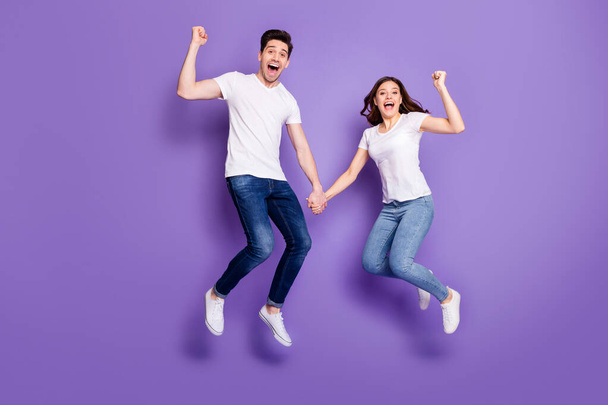 Full length photo pretty lady handsome guy couple jumping high up holding arms sporty people win race competitions wear casual t-shirts jeans pants shoes isolated purple color background - Fotó, kép