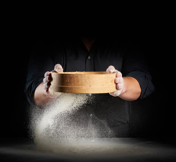 chef un hombre en un uniforme negro sostiene un tamiz de madera redonda en sus manos y tamiza harina de trigo blanco sobre un fondo negro, partículas vuelan en diferentes direcciones - Foto, imagen