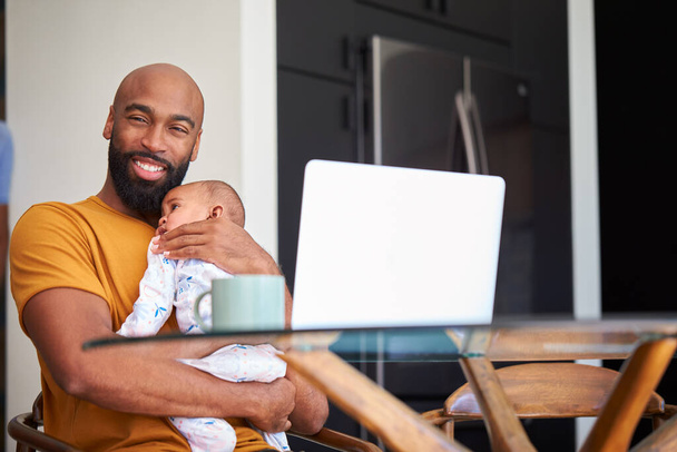 Evde gülümseyen Afrikalı Amerikalı baba, evde dizüstü bilgisayarla çalışırken bebeğe sarılıyor. - Fotoğraf, Görsel