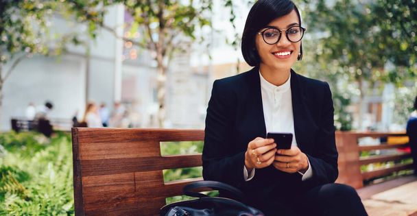 Happy successful businesswoman in formal wear and eyeglasses with cup of takeaway coffee sitting on wooden bench and browsing smartphone - Φωτογραφία, εικόνα