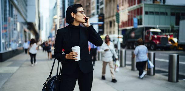 Confident young woman in elegant suit with cup of coffee talking on phone while standing in downtown against blurred exterior of modern buildings in New York City and looking away - Foto, afbeelding