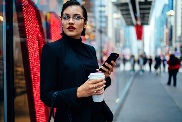 Femme à la mode en tenue chic debout près du mur de verre et smartphone de navigation tout en tenant une tasse de café et en regardant loin - Photo, image