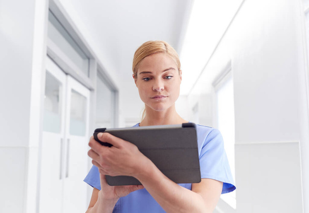 Female Doctor Wearing Scrubs In Hospital Corridor Using Digital Tablet - Фото, зображення