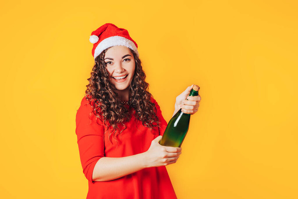 Mujer joven en vestido rojo y sombrero de Navidad abre una botella. - Foto, Imagen
