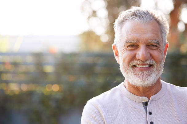Portrait Of Smiling Retired Senior Hispanic Man In Garden At Home Against Flaring Sun - Photo, image
