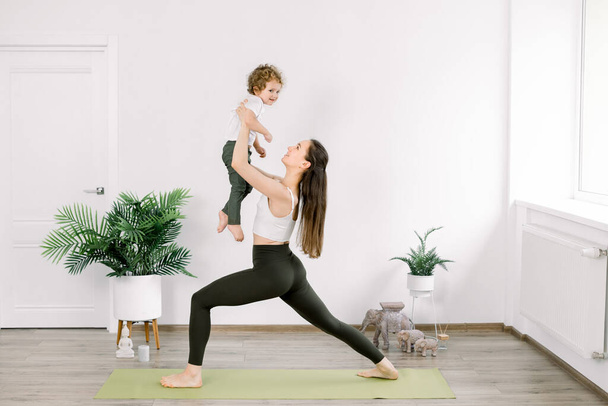 Mother and little son in sportswear doing yoga together at home. Beautiful young Caucasian mother practices yoga with her son, mom in yoga pose holds her child - Фото, изображение