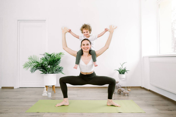 Beautiful young Caucasian woman and her little son are smiling while doing yoga together at home. Woman in yoga position with her son sitting on her shoulders - Photo, Image