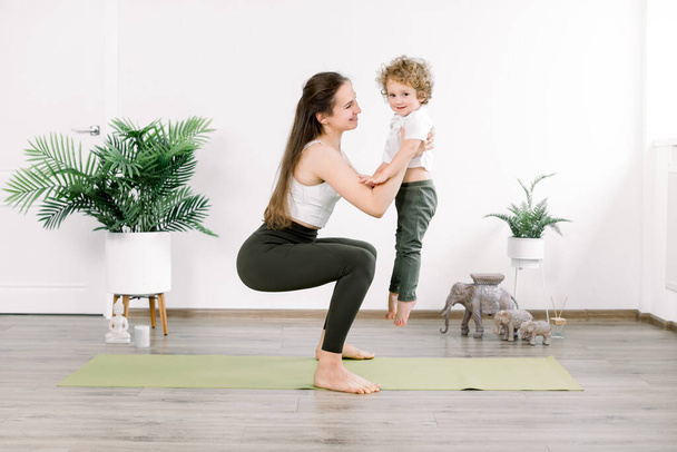 Pretty young Caucasian woman mother in sports wear does physical fitness exercises, squats on green mat, holding on hands her little kid son. Sport, yoga and motherhood concept - Foto, Imagem