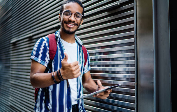 Half length portrait of dark skinned bearded male satisfied with application for navigation on digital tablet, smiling african american hipster guy gesturing showing thumb up looking at camera - Фото, изображение
