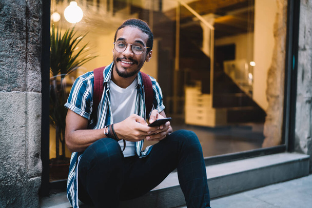 Handsome ethnic male in casual wear and eyeglasses sitting on stair near glass wall and messaging on social media on smartphone and looking at camera - Fotoğraf, Görsel