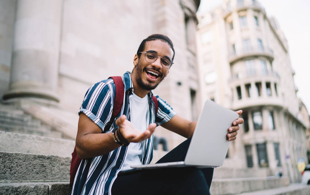 Carefree dark skinned hipster guy satisfied with learning online course outdoors having fun and laughing,positive african american millennial man gesture and looking at camera blogging and share media - Photo, image