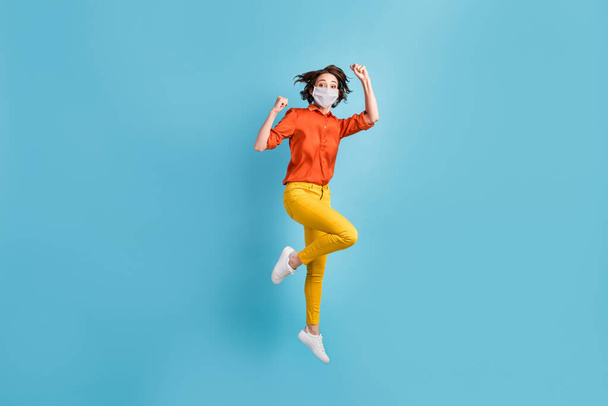 Full length photo of crazy lady jump fists rejoicing wear mask orange shirt yellow trousers sneakers isolated blue color background - Φωτογραφία, εικόνα