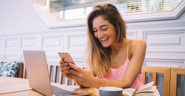 Весела усміхнена етнічна жінка-блогерка в повсякденному одязі, що працює над проектом онлайн через ноутбук та повідомлення на смартфоні, сидячи за столом у кафе
 - Фото, зображення
