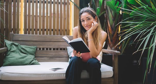 Happy female in casual clothes sitting on wooden bench and reading interesting book while resting on sunny terrace during weekend - Foto, immagini