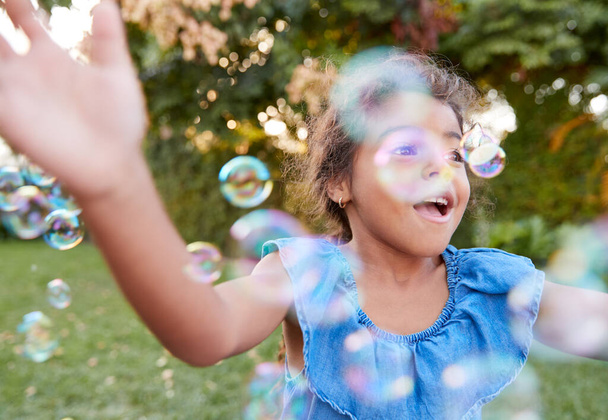 Young Hispanic Girl Chasing And Catching Bubbles In Garden - Foto, imagen