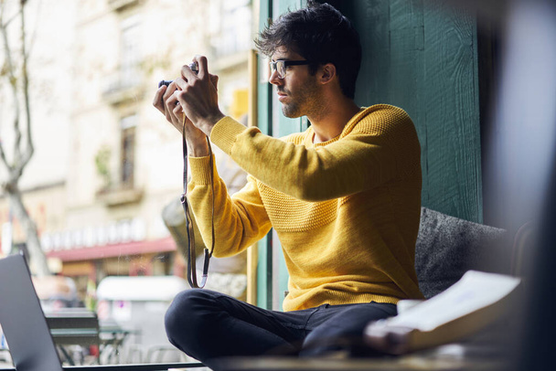 Pensive etnische man in casual outfit en bril zitten op vensterbank en het nemen van foto 's op fotocamera terwijl het hebben van pauze - Foto, afbeelding
