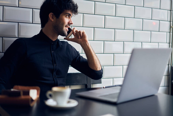 Positive ethnic male freelancer in casual wear smiling and looking away while having phone call during work on laptop in cafe - 写真・画像