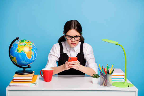 Frustrated girl college student sit table have geography globe lesson break mug beverage use smartphone lose race game wear white blouse black overall uniform isolated blue color background - Foto, Imagen