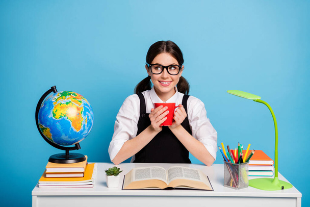 Photo of positive high school girl sit table hold beverage mug enjoy academic courses learning wear white blouse black overall uniform isolated over blue color background - 写真・画像