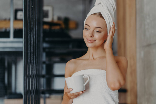 Photo of healthy thoughtful woman with healthy smooth skin drinks hot tea concentrated down, stands wrapped in white bath towel in cozy room, enjoys hygiene and beauty treatments, feels relaxed - Photo, Image