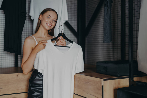 Positive smiling woman dresses for special occasion, holds white dress on hangers, poses over home wardrobe, buys clothes of latest trends, makes decision what to wear. Women, style, fashion concept - Photo, Image