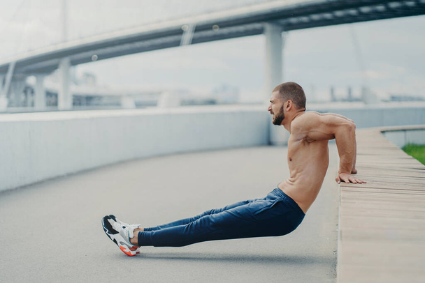 Sideways shot of muscular man does reverse push up exercise, trains arms, keeps perfect body and good physical shape, has active workout outdoor, wears sport trousers and sneakers, poses on bridge - Zdjęcie, obraz