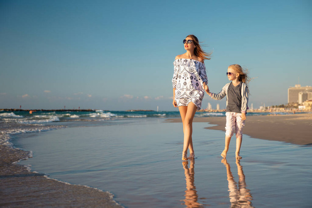 Küçük bir kızı olan mutlu bir anne kumsalda sörf yaparken yalınayak yürüyor. - Fotoğraf, Görsel