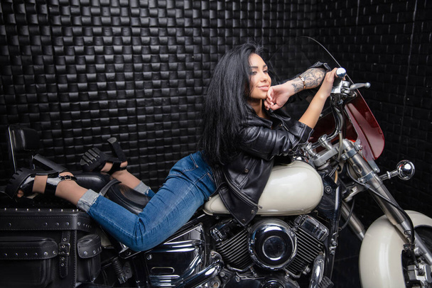 若い女性を魅了革のジャケットとジーンズを着たバイクに横たわっている - 写真・画像